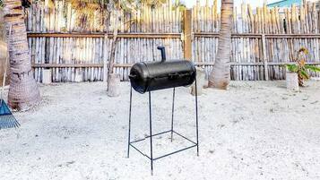 Un barbecue installé dans un espace extérieur sablonneux, agrémenté d’une clôture en bois et de palmiers.