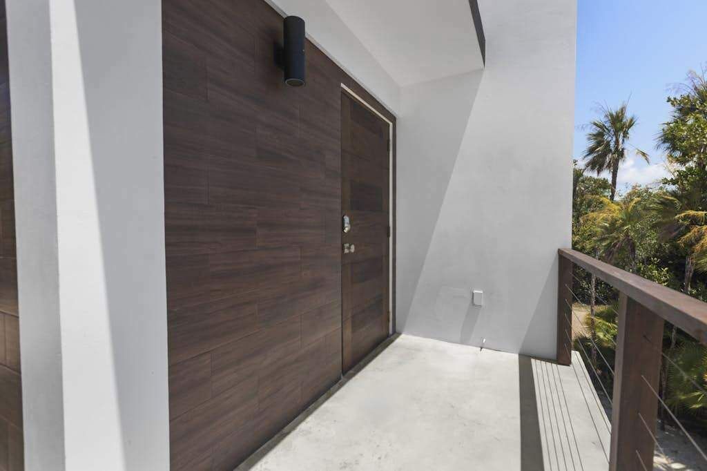 A balcony with a wooden floor, a dark wooden door, and a view of tropical vegetation.
