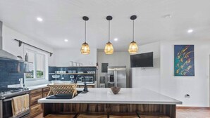 A modern kitchen with a central island, bar stools, pendant lighting, and a large TV on the wall.