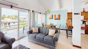 A living room with a grey sofa, a dining area with wicker chairs, a kitchen with wooden cabinets, and a balcony with a view of buildings. - Lake-view condo with reef-view balcony, washer & dryer, pool & central AC (San Pedro)
