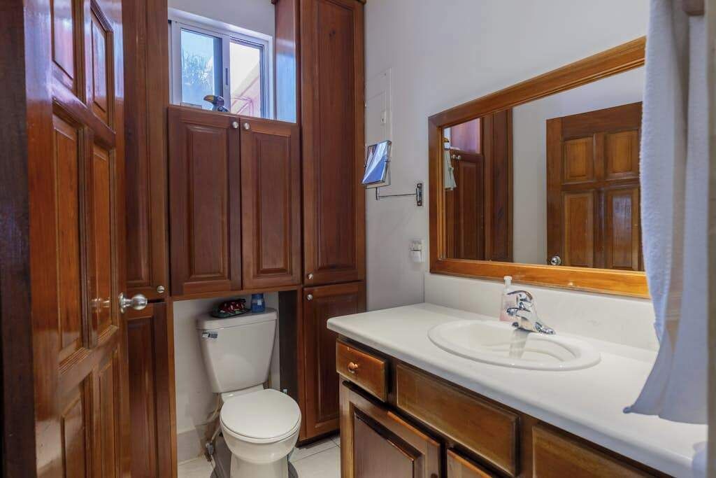 A bathroom with wooden cabinets, a white toilet, a sink with a mirror, and a window above the toilet.