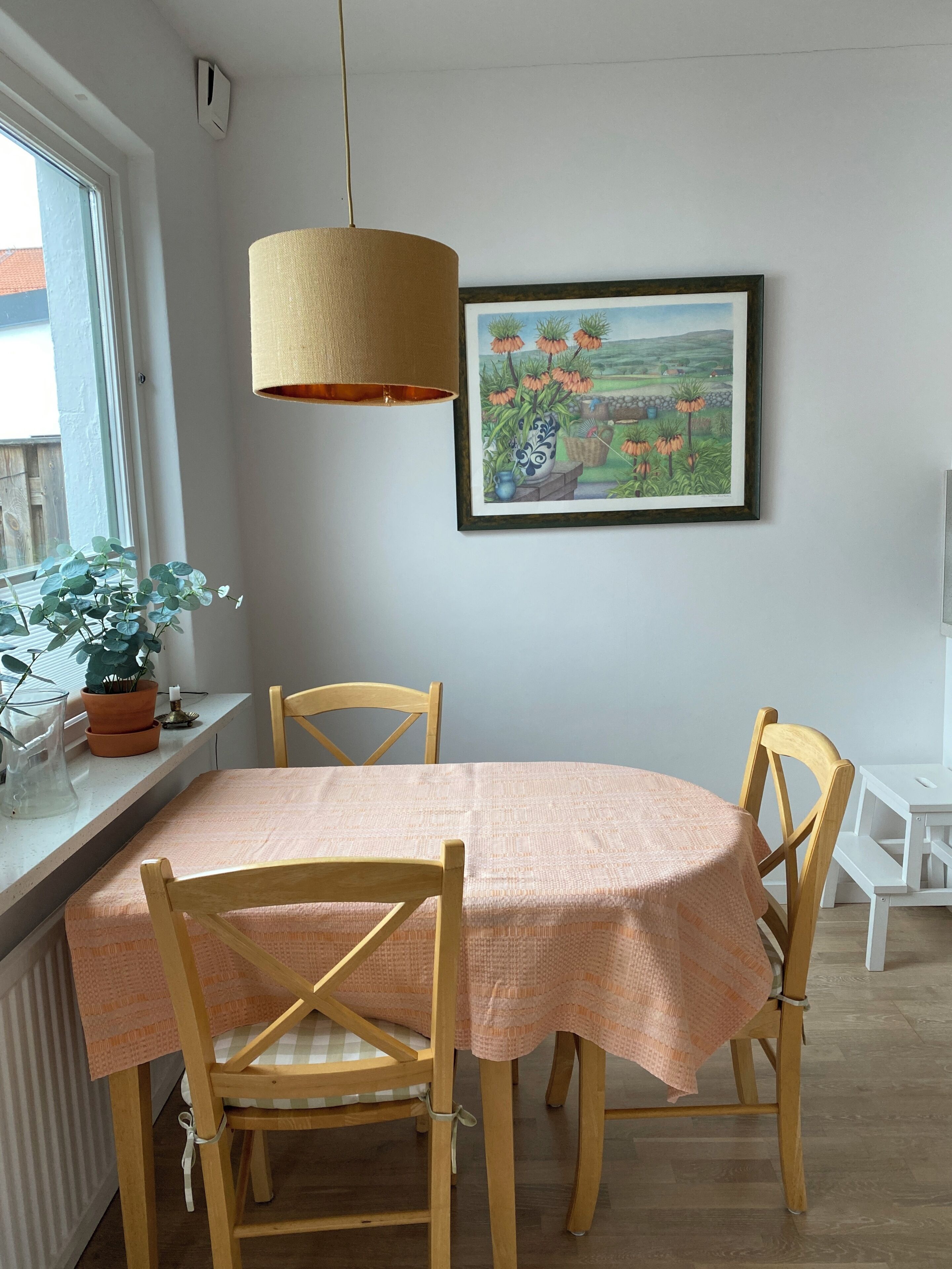 Ruokailutilassa on pyöreä pöytä, neljä puista tuolia, riippuvalaisin, ruukussa oleva kasvi ja seinällä oleva kehystetty maisemamaalaus.