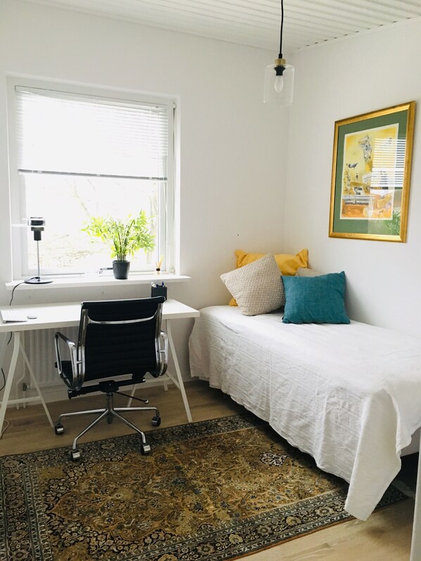 A bedroom with a bed, a desk, a chair, a window with blinds, a potted plant, and a framed picture on the wall.