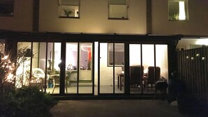 Un edificio residencial con un amplio acceso por puerta corrediza de vidrio, se ve un salón con muebles y un pequeño balcón con planta en maceta.