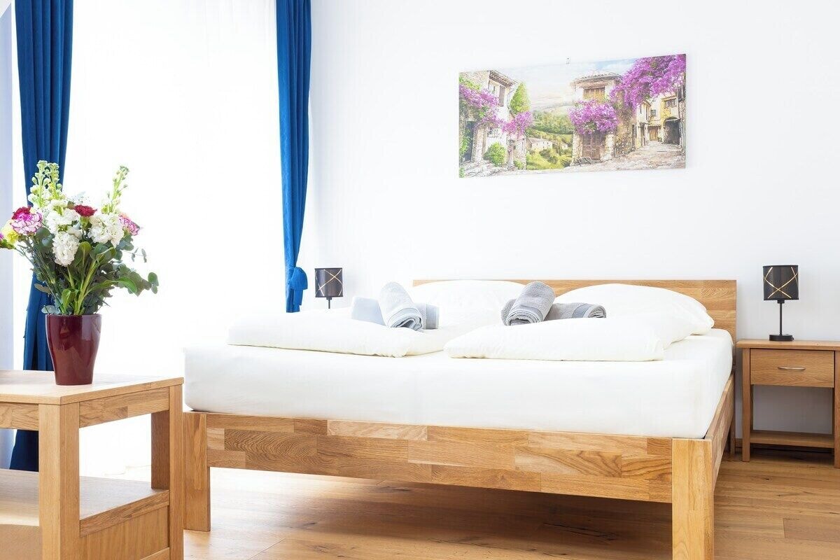 A bedroom with a wooden bed, a bedside table, a nightstand, a wall-mounted lamp, and a painting of a village scene.