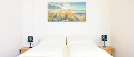 En pænt redt seng med hvide sengetøj, to sengeborde med lamper, og et ophængt maleri af en strandscene.