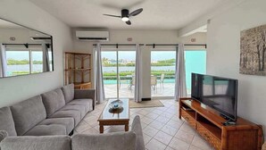 A living room with a sofa, a coffee table, a TV on a wooden stand, a ceiling fan, and large windows with a view of a body of water. - Escape to Paradise - Beautiful Lakeside Villa with pool access and WiFi (San Pedro)