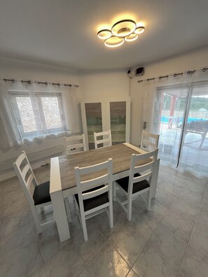 Een eetruimte met een houten tafel en witte stoelen, een plafondlamp en uitzicht door een glazen deur.