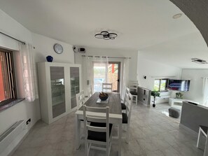A modern kitchen with white cabinetry, a dining area with a wooden table and chairs, and a TV mounted on the wall.