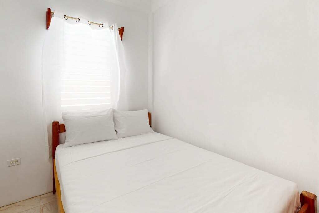 Uma cama de solteiro com roupa de cama branca, cabeceira de madeira e uma janela com cortina branca.
