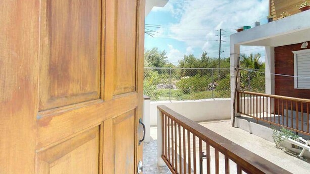 A wooden door leading to a balcony with a metal railing, a white wall, and a view of greenery and a clear sky. - Enchanting single-level living space with a mini kitchen in a prime area (Caye Caulker)