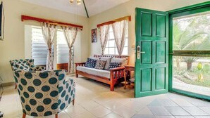 Una habitación con sofá, un sillón estampado, una puerta verde y vistas al exterior con vegetación.