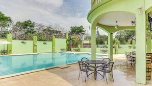Una piscina con un porche cubierto, mobiliario exterior y una estructura de edificio verde.