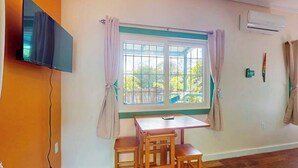 A room with a TV mounted on the wall, a window with curtains, a table and chairs, and an air conditioning unit. - Charming retreat with spacious outdoor areas, cozy interior & near to beach (Placencia)