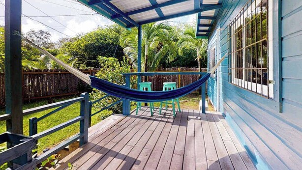A covered porch with a hammock, wooden deck, and a blue building with windows. - Charming retreat with spacious outdoor areas, cozy interior & near to beach (Placencia)
