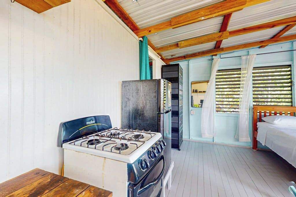 Une cuisine équipée d’une cuisinière à gaz, d’un four et d’un réfrigérateur, une table en bois et un lit avec du linge de lit blanc.