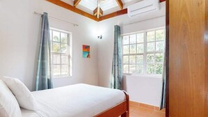 Een slaapkamer met een bed, twee ramen met gordijnen, een plafondventilator en een aan de muur bevestigde airconditioner.