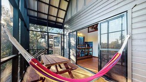 A hammock on a wooden deck with a table, large windows, and a view of trees.