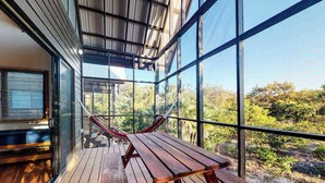 A wooden deck with a table and chairs, large windows, and a view of greenery.