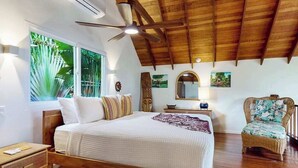 A bedroom with a wooden ceiling, a bed with white bedding, a nightstand, a chair, and a window with a view of greenery. - Romantic beachfront home w ocean views, dock, hammock, veranda & free WiFi (Placencia)
