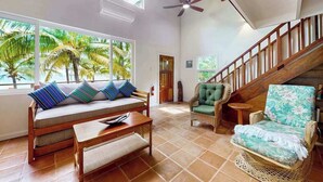 A spacious living area with a sofa, chairs, a wooden table, a staircase, and large windows with a view of palm trees. - Romantic beachfront home w ocean views, dock, hammock, veranda & free WiFi (Placencia)