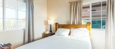 Habitación de hotel con cama, mesita de noche, lámpara y dos ventanas con cortinas.