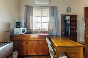 Une petite cuisine équipée d’un four à micro-ondes, d’une machine à café et d’un évier. On y trouve une table et une chaise en bois, ainsi qu’une fenêtre avec des rideaux.