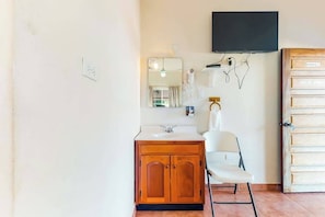 Une salle de bain avec un meuble en bois, une chaise blanche, un miroir, un téléviseur fixé au mur et une porte sur laquelle est apposée une pancarte « Interdiction de fumer ».
