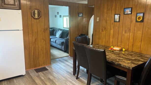 Ein Zimmer mit Holzwänden, einem Esstisch mit Stühlen, einem Kühlschrank und einer Couch im Hintergrund.