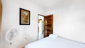Une chambre à coucher avec un lit, un ventilateur sur pied, une porte en bois et un tableau encadré accroché au mur.