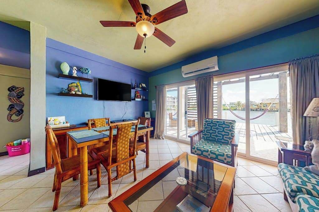 Una sala de estar con una mesa de comedor, sillas, un sofá, un ventilador de techo, un televisor y un ventanal grande.