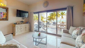 A living room with a sofa, a coffee table, a TV, a cabinet, and a sliding glass door leading to an outdoor area with a bicycle and a table. - Beachfront condo with pool, amazing ocean view, & W/D - near restaurants (San Pedro)