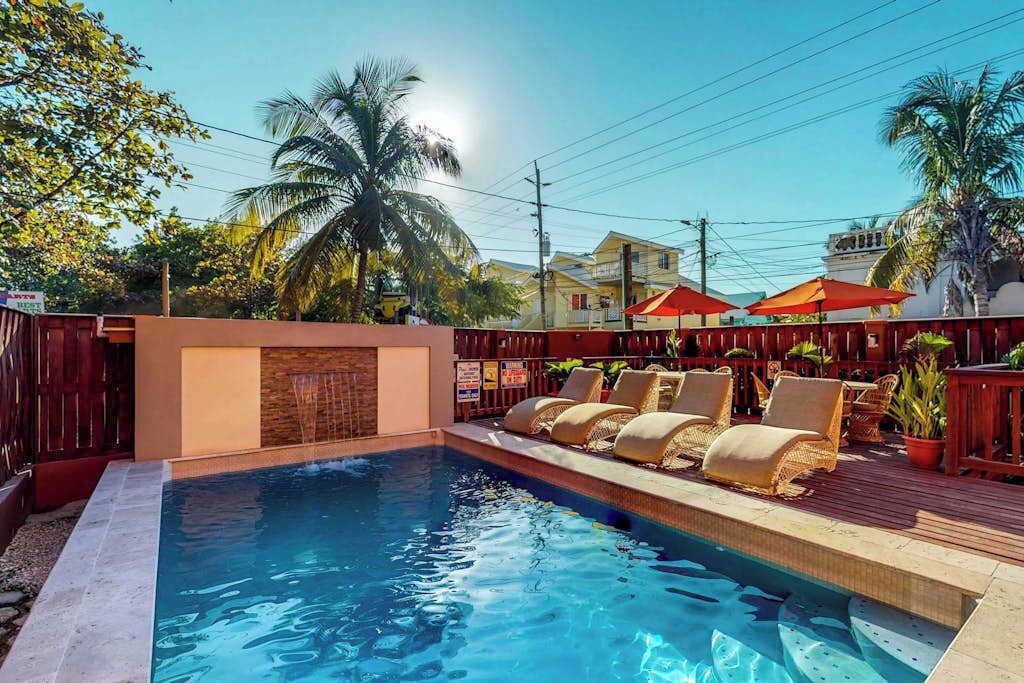 Ein Swimmingpool mit Liegestühlen und Sonnenschirmen, umgeben von Palmen und Gebäuden.