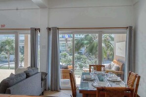 A dining area with a table set for four, a sofa, and a view of palm trees through large windows. - Ground-level condo w WiFi, central AC, & pool - walk to the beach (San Pedro)