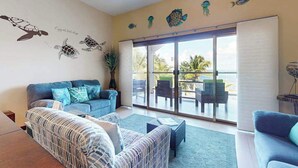 A living room with a sofa, a patterned armchair, a coffee table, and a sliding glass door leading to an outdoor seating area with chairs and a table. - Oceanfront condo with pool, gorgeous ocean views, WiFi & partial AC (San Pedro)