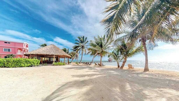 Une plage avec un abri à toit de chaume, des palmiers et un bâtiment rose.