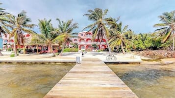 A wooden dock leading to a building with a red facade and white balconies, surrounded by palm trees.