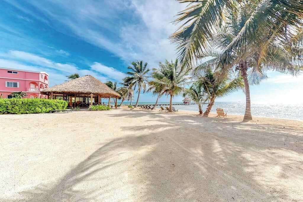 Uma praia com uma cabana de telhado de palha, palmeiras e um prédio rosa.