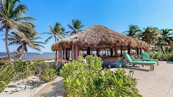 A beachside hut with a thatched roof, surrounded by palm trees and a lounge area with chairs.