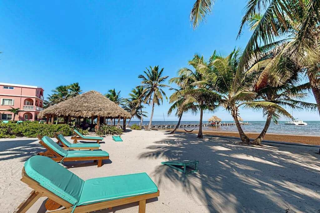Ein Strand mit Liegestühlen, Palmen, einer mit Stroh gedeckten Struktur und einem Gebäude im Hintergrund.
