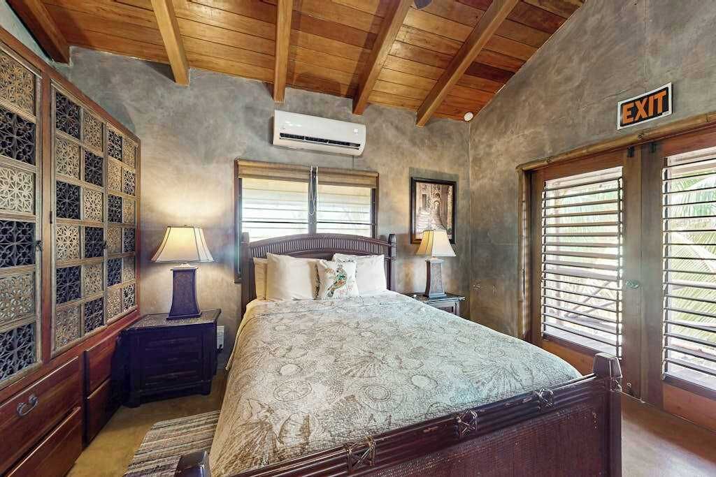 Un dormitorio con cama, dos mesitas de noche, equipo de aire acondicionado, ventana con persianas y señal de salida.