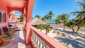 A pink building with a balcony overlooking a beach with palm trees and a thatched-roof structure. - Beachfront condo with beach access, dock, pool, balcony, & ocean views (San Pedro)