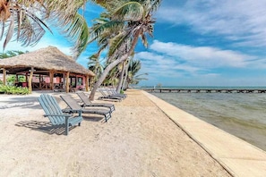 Ein Strand mit Liegestühlen, einer Struktur mit Reetdach, Palmen und ruhiger See.