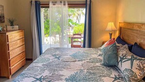 Une chambre à coucher comprenant un lit, une table de chevet, une lampe, une commode en bois et une vue sur l’extérieur par une fenêtre ornée de rideaux.