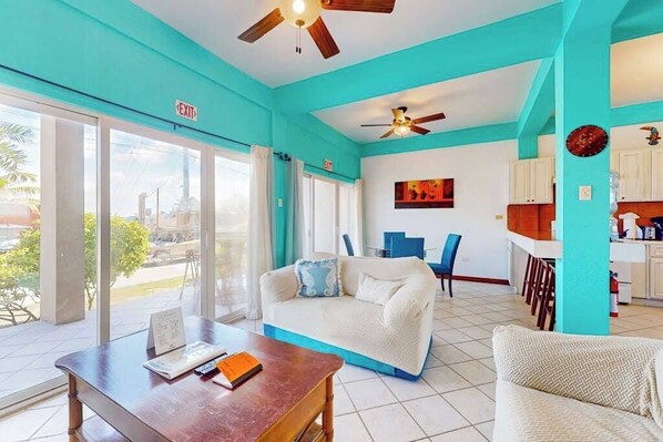 A room with a white sofa, a wooden table, a ceiling fan, a kitchen area, and an exit sign. - Lagoon front apartment w shared pool - two minutes from the beach (San Pedro)