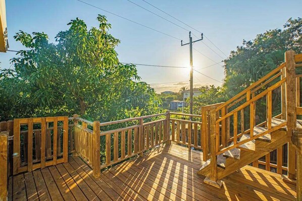 A wooden deck with a railing, surrounded by greenery and a clear sky. - Spacious coastal house with outdoor pool, deck & near beach (Placencia)