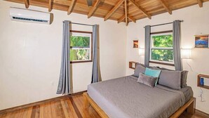 Una camera da letto con un letto di legno, due finestre con tende, un condizionatore d'aria e un soffitto in legno.