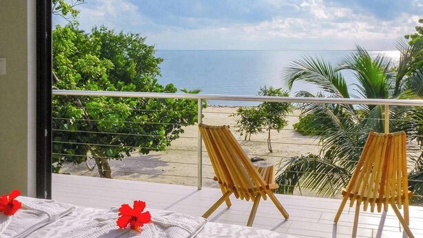Un balcón con vista a la playa, dos sillas de madera y toallas con flores rojas.