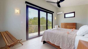 Un dormitorio con cama, un banco de madera, un ventilador de techo y vistas a través de un ventanal corredero.