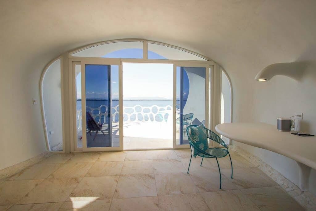 A room with a view of the sea, a curved ceiling, a glass door, a chair, and a white counter.